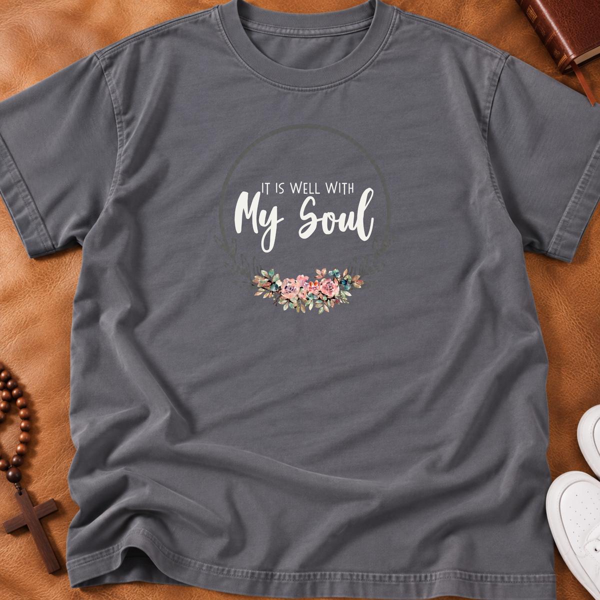 A storm colored short-sleeve graphic T-shirt laid flat on a white wood background, styled with blue jeans, a book, and a beaded bracelet. The shirt has a circular floral print with the text 'It Is Well With My Soul.