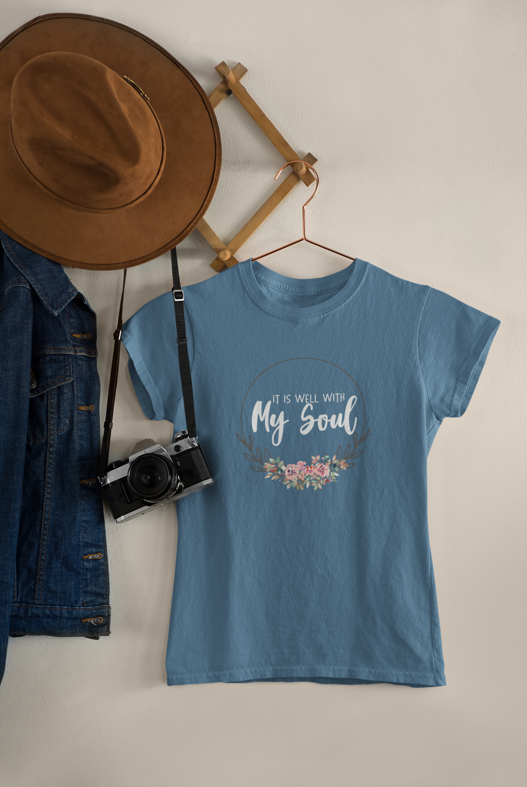 A light blue graphic bible faith t-shirt by SeedSowerGraphics hanging on a wooden hanger against a warm beige wall, styled with a brown wide-brim hat, a denim jacket, and a vintage camera on a wall-mounted wooden rack.