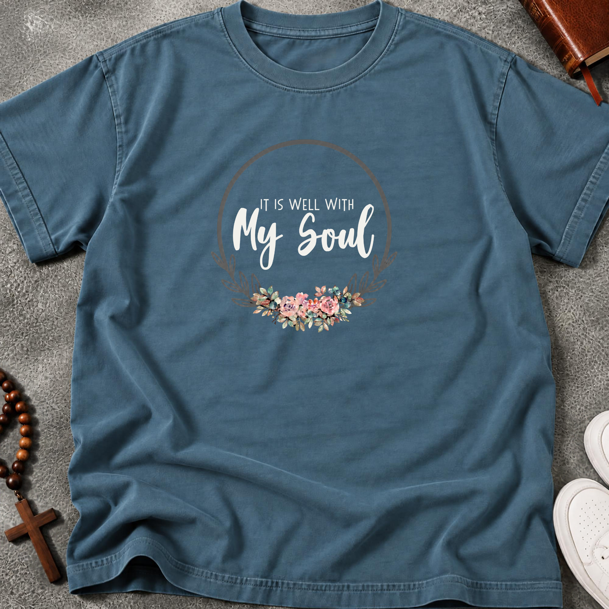 A slate blue graphic t-shirt laid flat on a gray textured surface, featuring a circular floral design with the text 'IT IS WELL WITH My Soul'.