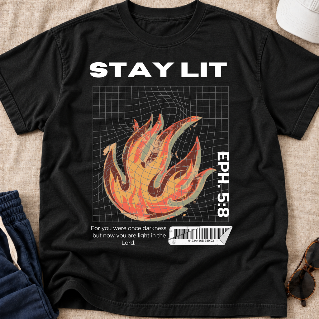 A black graphic t-shirt laid flat on a beige fabric surface, featuring the words 'STAY LIT' in white above a stylized orange and yellow flame over a grid, with 'EPH. 5:8' and a small quote printed on the front.