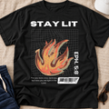A black graphic t-shirt laid flat on a beige fabric surface, featuring the words 'STAY LIT' in white above a stylized orange and yellow flame over a grid, with 'EPH. 5:8' and a small quote printed on the front.