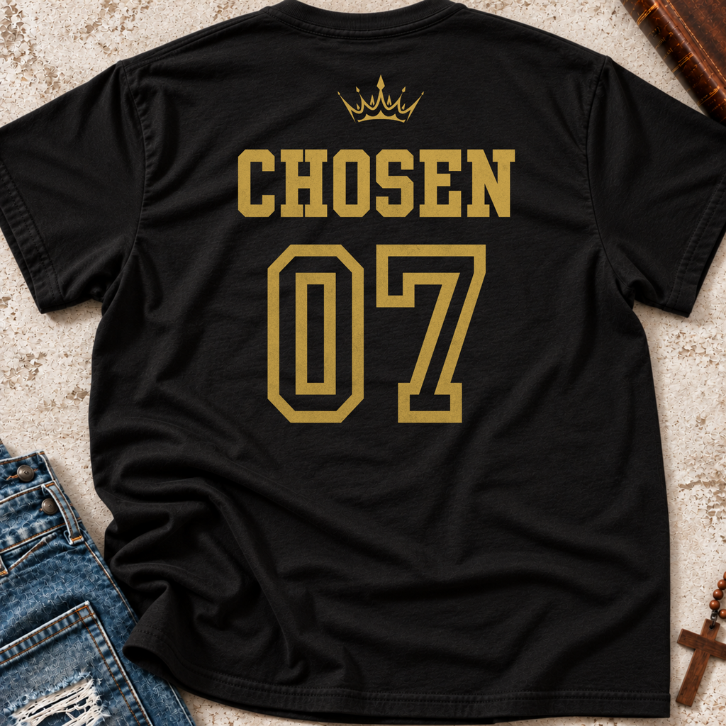 A black short-sleeve T-shirt laid flat on a textured beige surface, showing the back with the word 'CHOSEN' in gold above the number '07' in a sports-style outline, plus a small gold crown graphic. Blue jeans and a wooden book are partially visible as styling props.