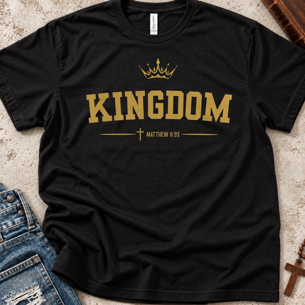 A black short-sleeve T-shirt laid flat on a textured light surface, featuring a gold crown graphic and the word 'KINGDOM' with 'MATTHEW 6:33' and a cross beneath. Blue jeans and a wooden book are partially visible as styling props.