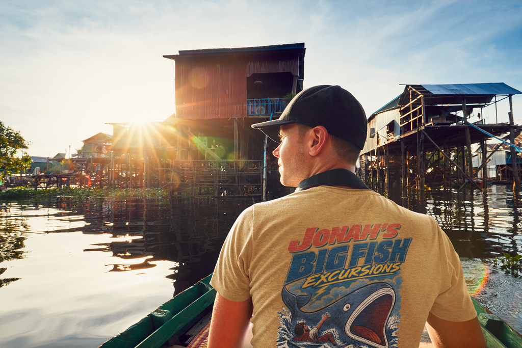 A man wearing a goldish Christian graphic t-shirt by SeedSowerGraphics with a colorful 'Jonah's Big Fish Excursions' graphic on the back, standing in a boat and looking at a sunset over a lake with stilted houses.