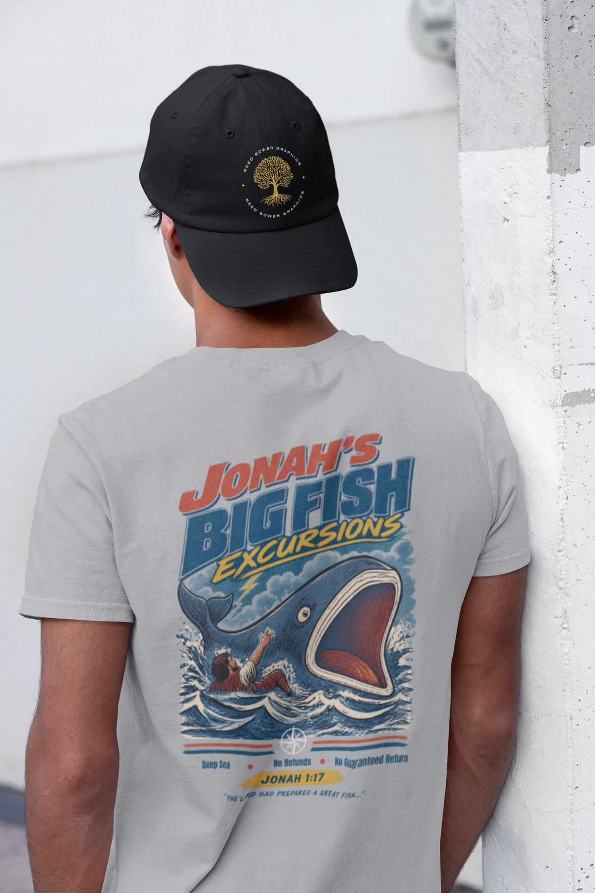 A man viewed from behind wearing a light gray Christian graphic t-shirt by SeedSowerGraphics with a large graphic print of a blue whale jumping out of the water, with the text 'JONAH'S BIG FISH Excursions'. He is also wearing a black baseball cap with a gold tree logo.