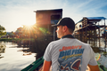 A man wearing a black baseball cap and a light gray Christian graphic t-shirt by SeedSowerGraphics with a 'Jonah's Big Fish Excursions' graphic on the back, seen from behind as he looks out of a boat on a river with stilted houses in the background during sunset.