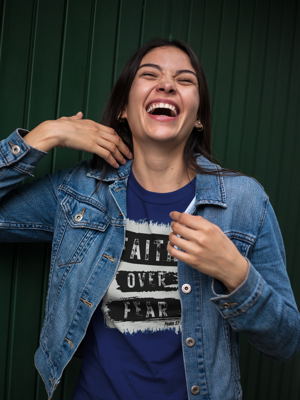 A woman laughing joyfully while wearing a blue denim jacket over a navy blue Christian graphic t-shirt that reads 'FAITH OVER FEAR' in white and black. She is posing in front of a dark green corrugated metal wall.