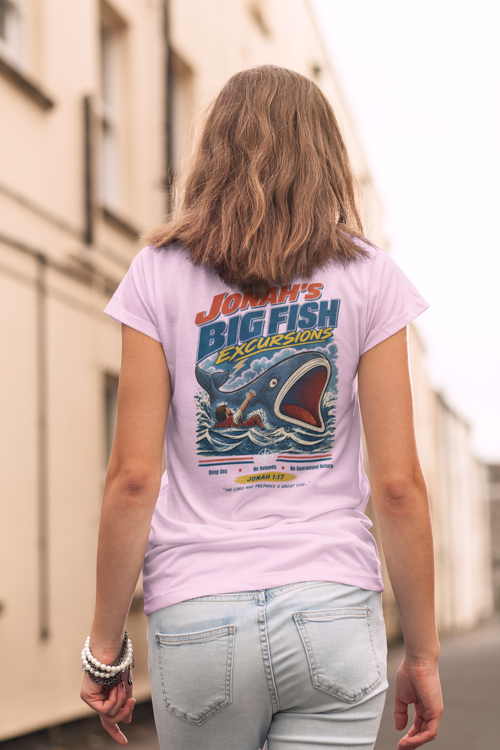 A girl viewed from behind wearing a light pink Christian graphic t-shirt by SeedSowerGraphics with a 'Jonah's Big Fish Excursions' design, paired with light-wash jeans in an outdoor street setting.