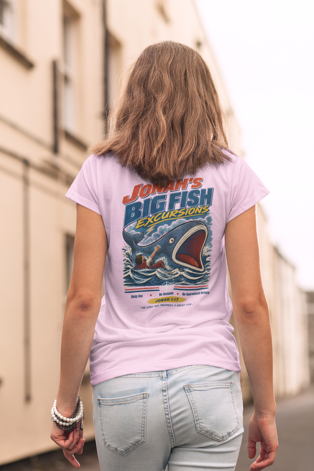 A girl viewed from behind wearing a light pink Christian graphic t-shirt by SeedSowerGraphics with a 'Jonah's Big Fish Excursions' design, paired with light-wash jeans in an outdoor street setting.