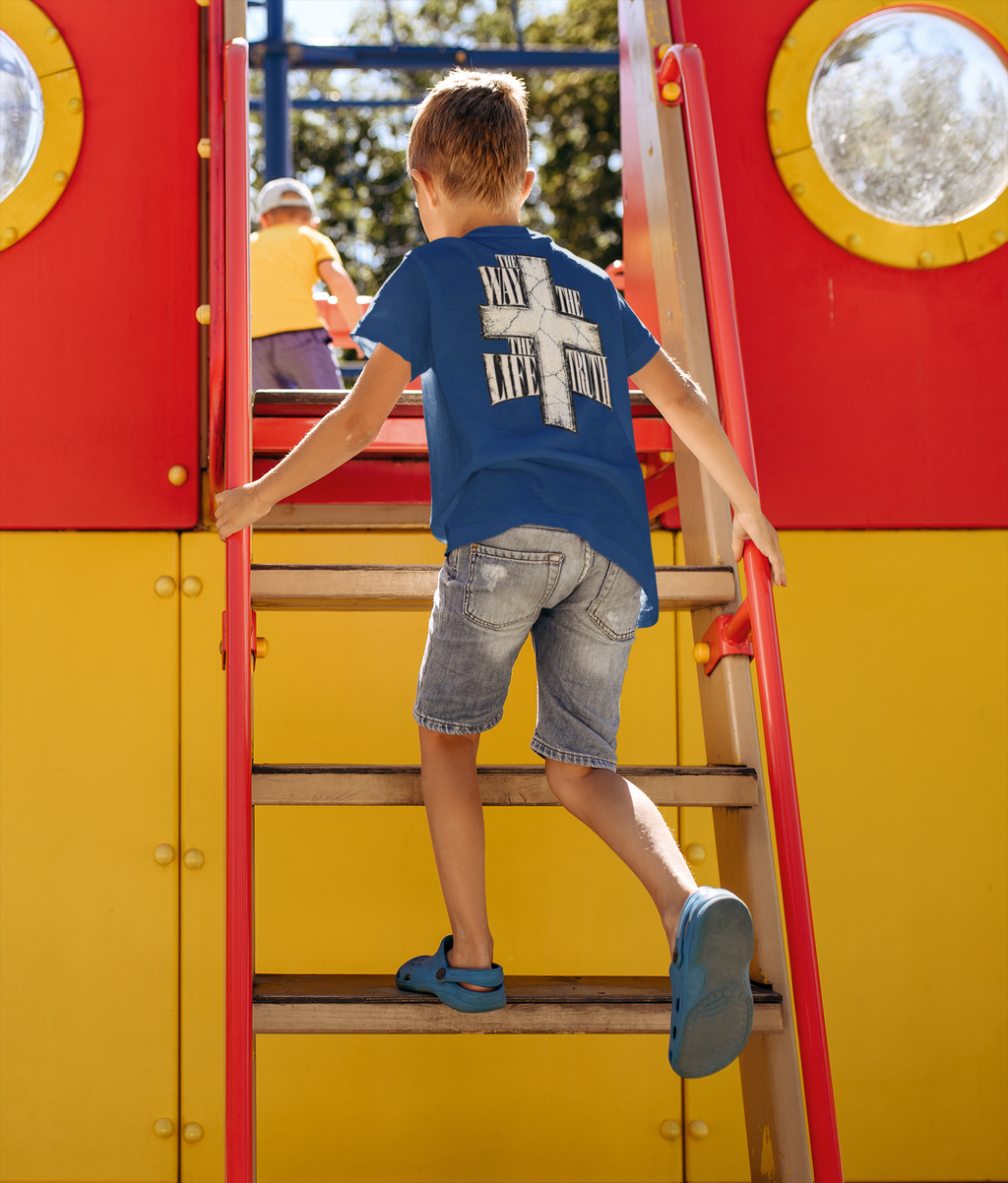 A young boy wearing a blue t-shirt with a white cross and the text 'HIS IS THE LIFE OF IT' on the back, denim shorts, and blue sandals is climbing a wooden ladder on a bright red and yellow playground structure.