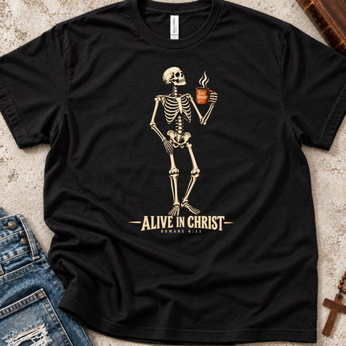 A black short-sleeve graphic T-shirt laid flat on a textured light surface, with a cream skeleton holding a coffee mug and the text 'ALIVE IN CHRIST ROMANS 8:11' printed below. Blue jeans and small decorative props are partially visible around the shirt.