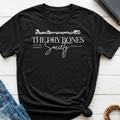 A black short-sleeve graphic T-shirt laid flat on a white wood surface, styled with blue jeans, a notebook, and a beaded bracelet nearby. The shirt has a white printed design reading “THE DRY BONES Society” with smaller text and decorative skull-and-flesh details above.