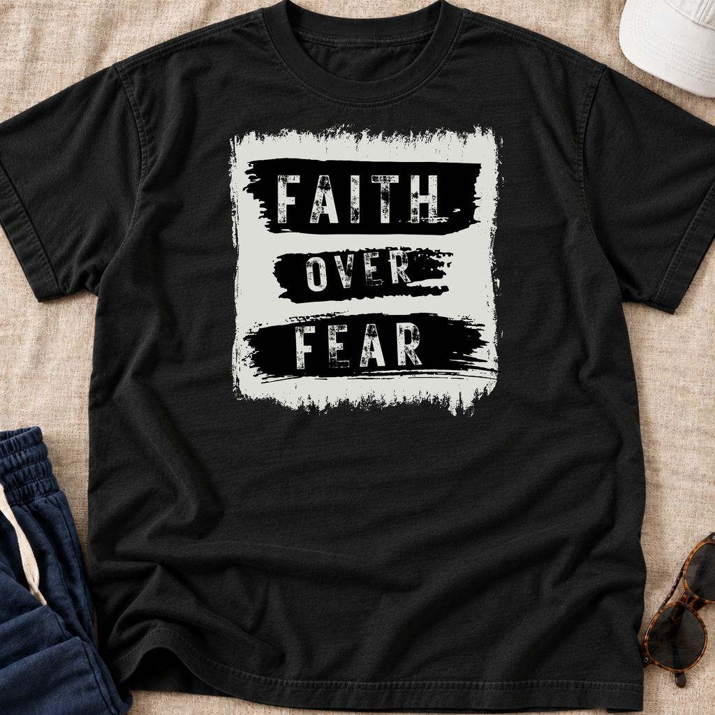 A black short-sleeve t-shirt laid flat on a beige fabric surface, featuring a distressed white square graphic with the words 'FAITH OVER FEAR' in black. Partially visible props include blue jeans, a white cap, and sunglasses around the edges.