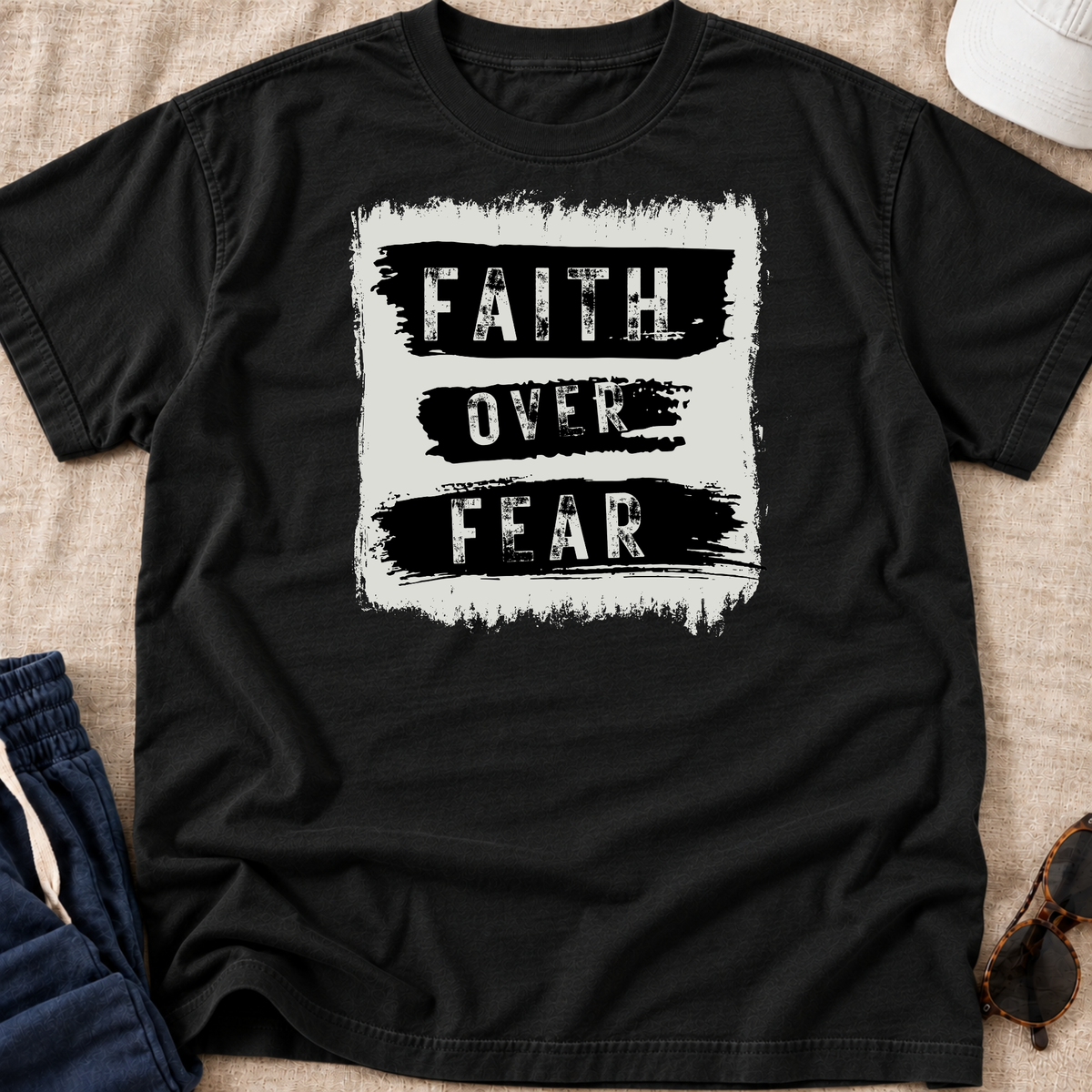 A black short-sleeve t-shirt laid flat on a beige fabric surface, featuring a distressed white square graphic with the words 'FAITH OVER FEAR' in black. Partially visible props include blue jeans, a white cap, and sunglasses around the edges.
