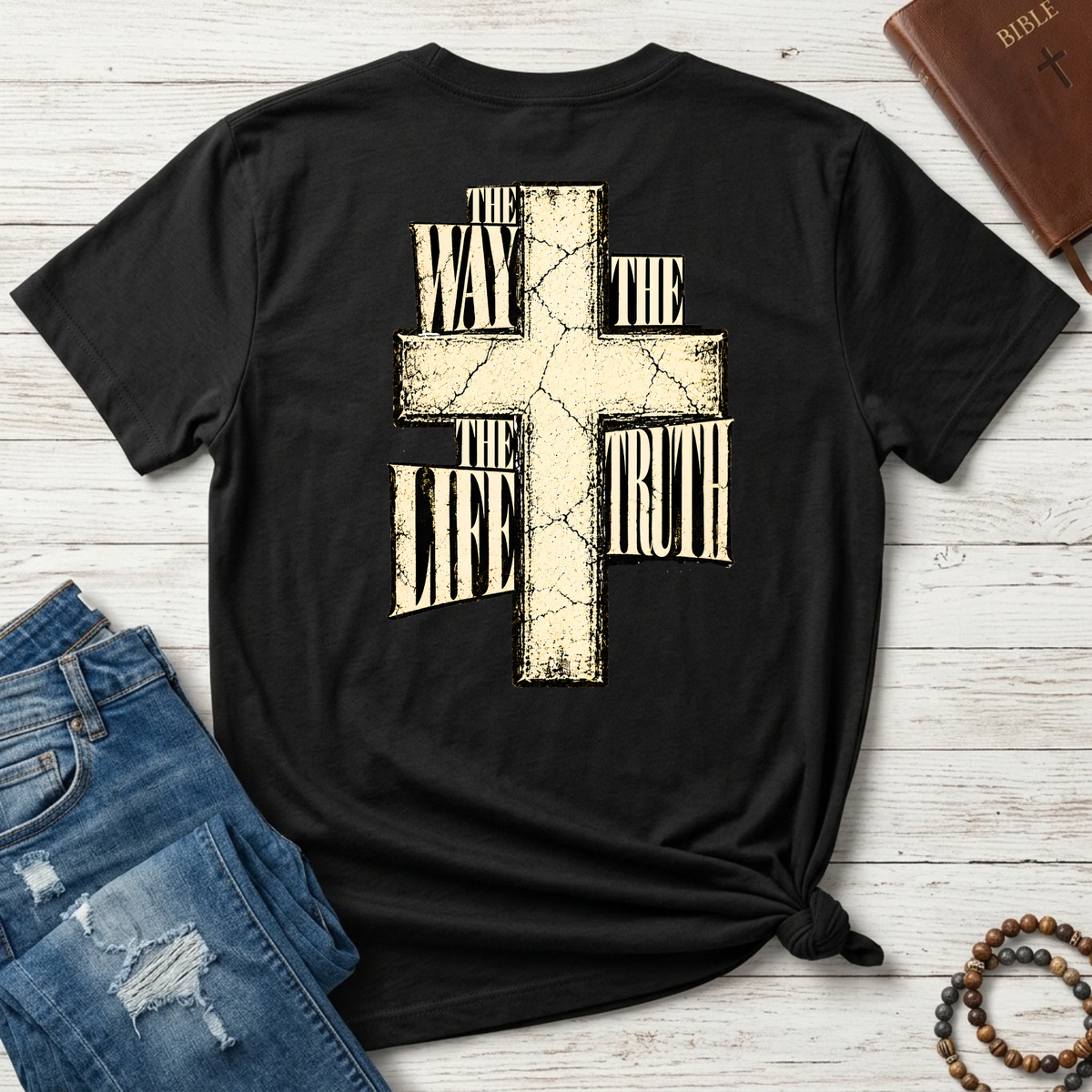 A black short-sleeve t-shirt with a distressed beige cross graphic on the back, featuring the text 'THE WAY THE LIFE THE TRUTH' integrated into the cross. The shirt is styled in a flat-lay arrangement on a white wood background, surrounded by a brown leather Bible, blue jeans, and a wooden bead rosary.