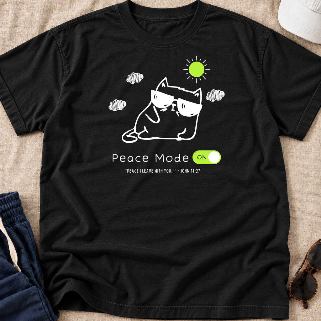 A black short-sleeve graphic T-shirt laid flat on a beige fabric surface, with a white line drawing of a cat wearing sunglasses, a green sun, clouds, and the text 'Peace Mode ON' plus a small scripture quote. Partial jeans, sunglasses, and a hat are visible around the shirt as styling props.