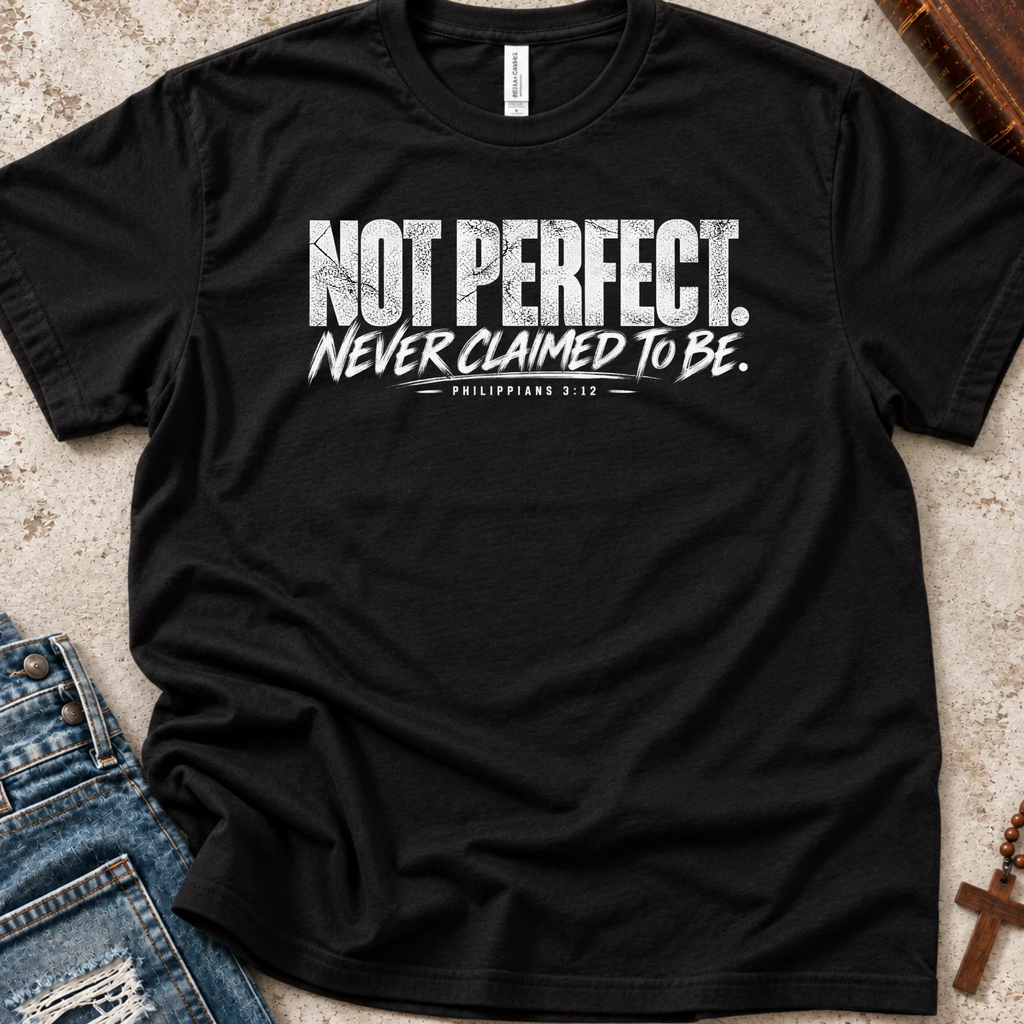 A black short-sleeve graphic T-shirt laid flat on a textured light surface, styled with blue jeans, a book, and a cross. The shirt has a distressed white text print reading “NOT PERFECT. Never Claimed to Be