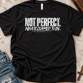 A black short-sleeve graphic T-shirt laid flat on a textured light surface, styled with blue jeans, a book, and a cross. The shirt has a distressed white text print reading “NOT PERFECT. Never Claimed to Be