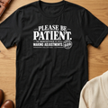 A black short-sleeve graphic T-shirt laid flat on a wooden surface, with white text reading 'PLEASE BE PATIENT. THE MANUFACTURER IS STILL MAKING ADJUSTMENTS' and a circular badge that says 'BE GOD.