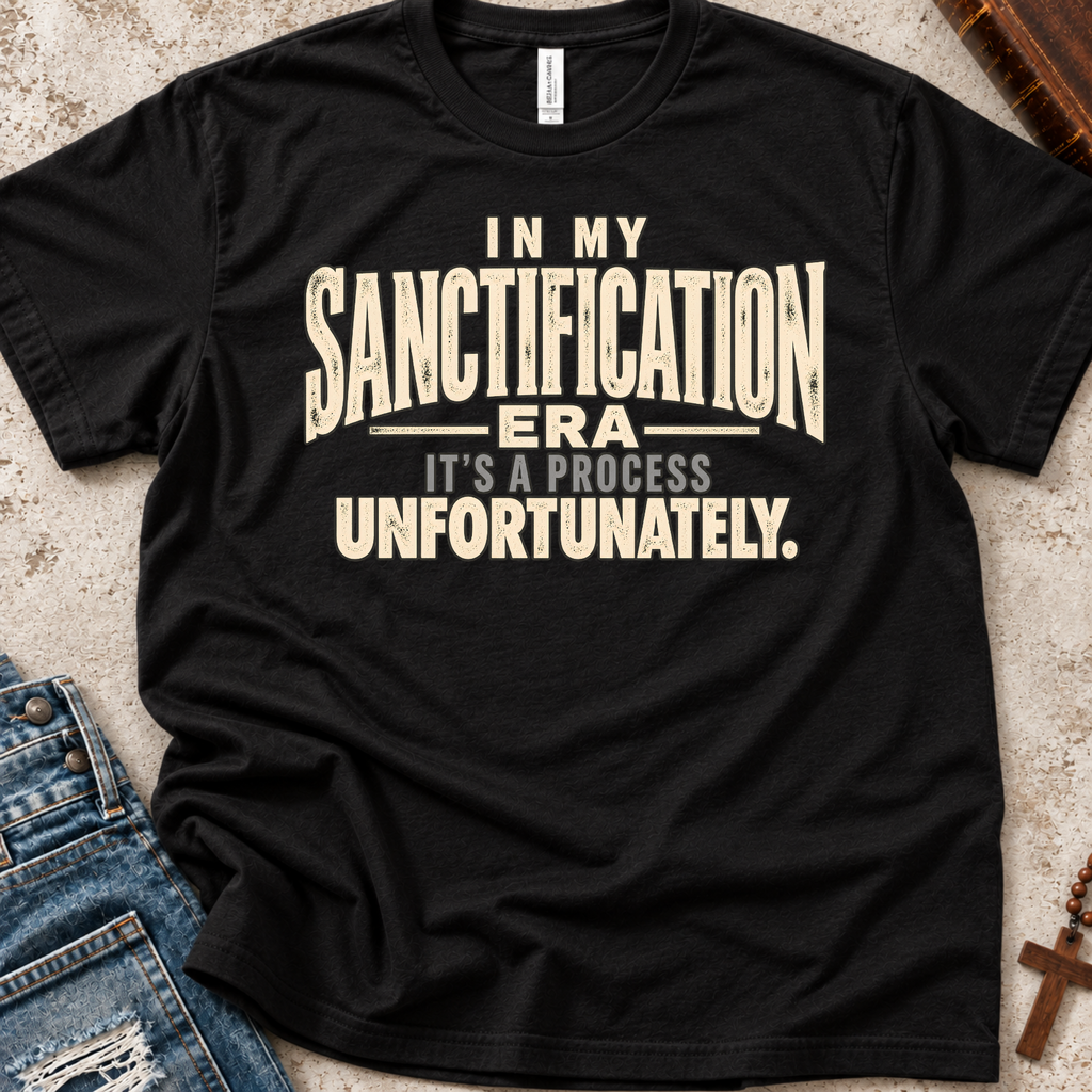 A black short-sleeve graphic T-shirt laid flat on a textured light surface, with blue jeans partially visible at the lower left and a wooden cross at the lower right. The shirt has large distressed cream text reading, "IN MY SANCTIFICATION ERA IT'S A PROCESS UNFORTUNATELY.