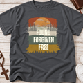 A dark gray crewneck Christian t-shirt with a retro-style graphic print featuring a sunset, pine trees, and the text 'FOUND FORGIVEN FREE' in white. The shirt is displayed in a flat-lay arrangement on a light wood background with a rosary, a book, and white sneakers as props.