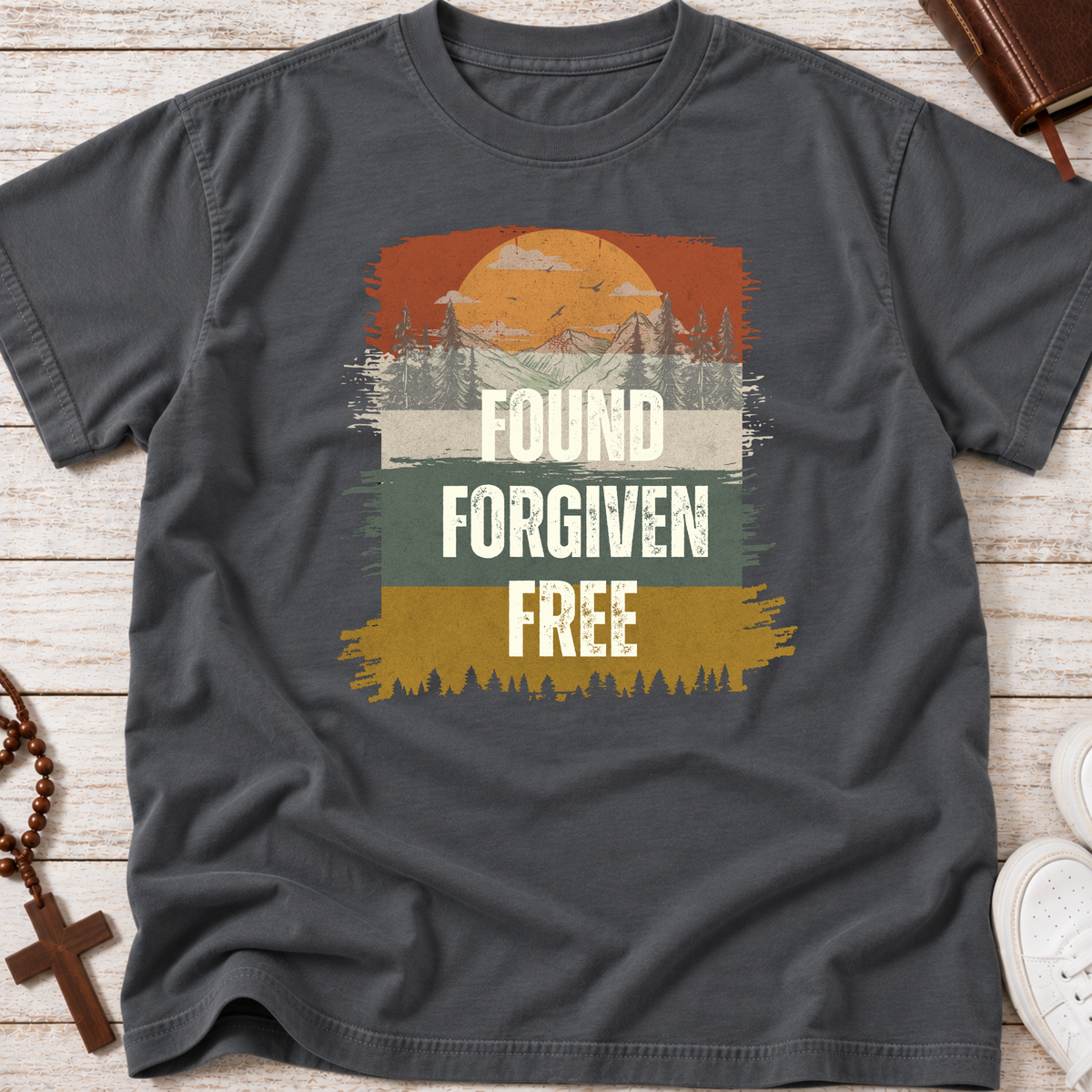 A dark gray crewneck Christian t-shirt with a retro-style graphic print featuring a sunset, pine trees, and the text 'FOUND FORGIVEN FREE' in white. The shirt is displayed in a flat-lay arrangement on a light wood background with a rosary, a book, and white sneakers as props.