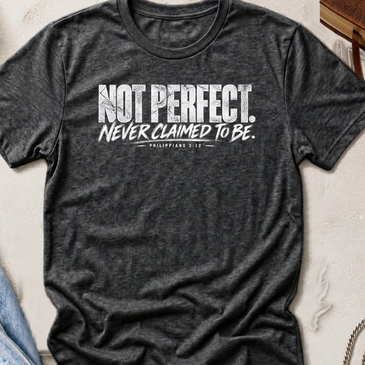 A dark heather gray graphic t-shirt laid flat on a textured light beige surface. The shirt features a distressed white text print that reads 'NOT PERFECT.