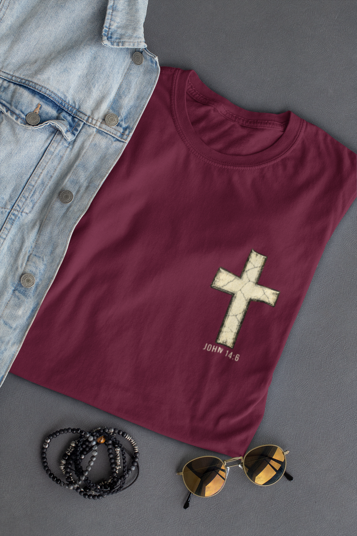 A folded maroon Christian graphic t-shirt with a distressed white cross and 'JOHN 14:6' printed on the front, styled in a flat lay with a light blue denim shirt, black beaded bracelets, and sunglasses on a dark gray background.