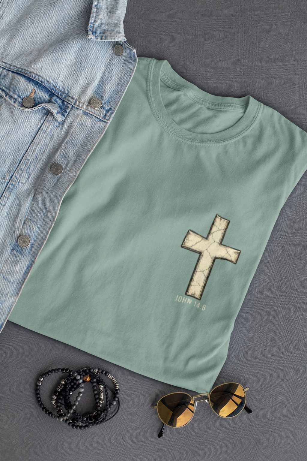 A sage green Christian t-shirt by SeedSowerGraphics with a black and white crown graphic and the text 'John 1:1' printed on the chest, styled in a flat-lay with a denim jacket, black beaded bracelets, and sunglasses on a dark gray background.