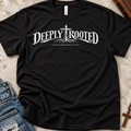A black short-sleeve Christian graphic T-by SeedSowerGraphics shirt laid flat on a textured light surface, with blue jeans and a wooden cross partially visible as styling props. The shirt has a large white 'DEEPLY ROOTED' graphic across the chest with a cross and the words 'COLOSSIANS 2:7' beneath it.