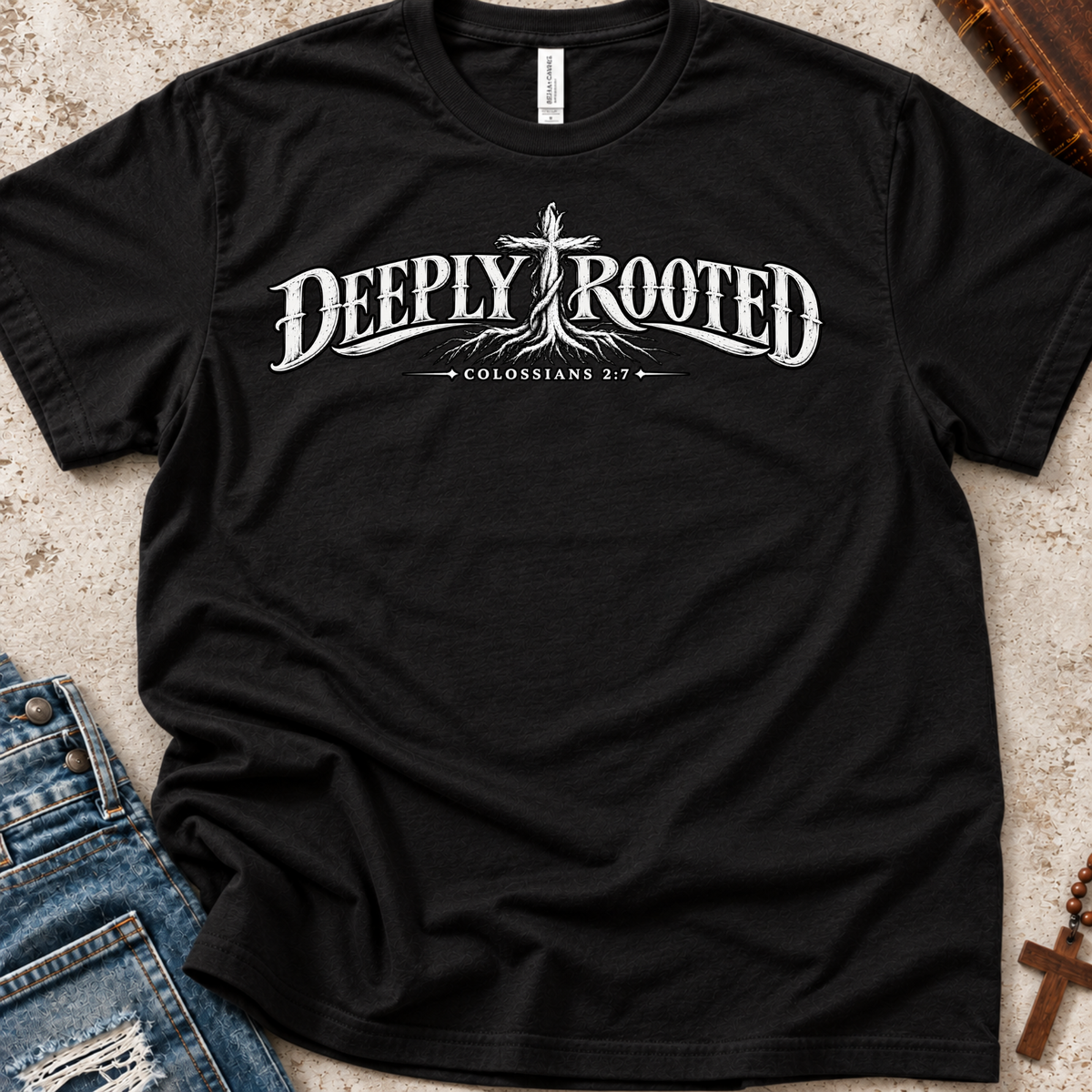 A black short-sleeve Christian graphic T-by SeedSowerGraphics shirt laid flat on a textured light surface, with blue jeans and a wooden cross partially visible as styling props. The shirt has a large white 'DEEPLY ROOTED' graphic across the chest with a cross and the words 'COLOSSIANS 2:7' beneath it.