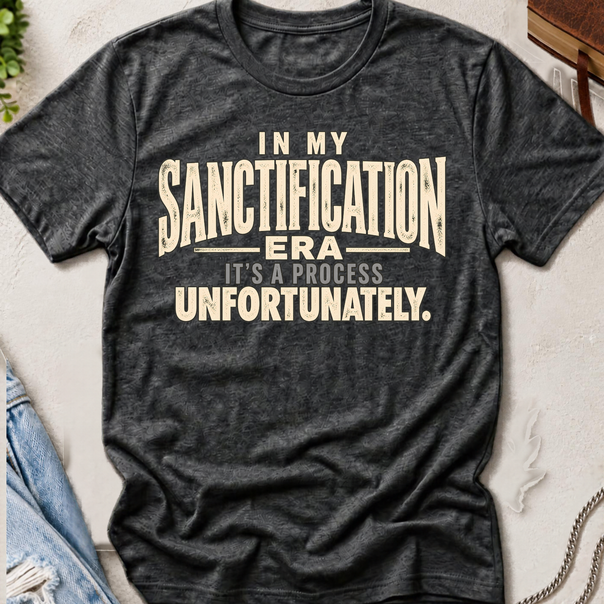 A dark heather gray graphic t-shirt laid flat on a textured light background, featuring the text 'IN MY SANCTIFICATION ERA IT'S A PROCESS UNFORTUNATELY.' in cream-colored distressed lettering.