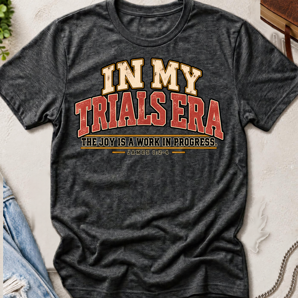 Heather grey christian t-shirt by SeedSowerGraphics  laid out amongst props with typography that says "in my trials era' the joy is a work in progress. James 1:2-4 in cream, red, and black lettering