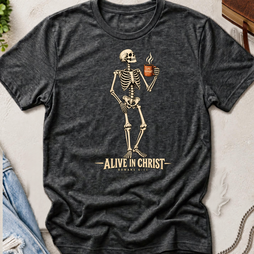 A dark heather gray graphic t-shirt laid flat on a textured light background, featuring a beige skeleton holding a coffee mug with the text 'ALIVE IN CHRIST ROMANS 8:6' printed below.