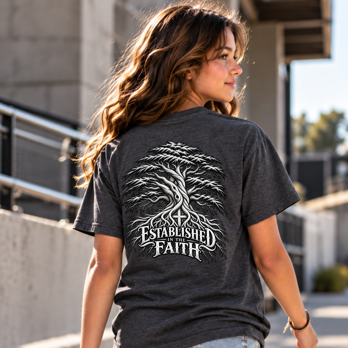 A woman wearing a dark heather gray t-shirt with a large white graphic of a tree and the text 'ESTABLISHED IN FAITH' on the back, shown in an outdoor setting.