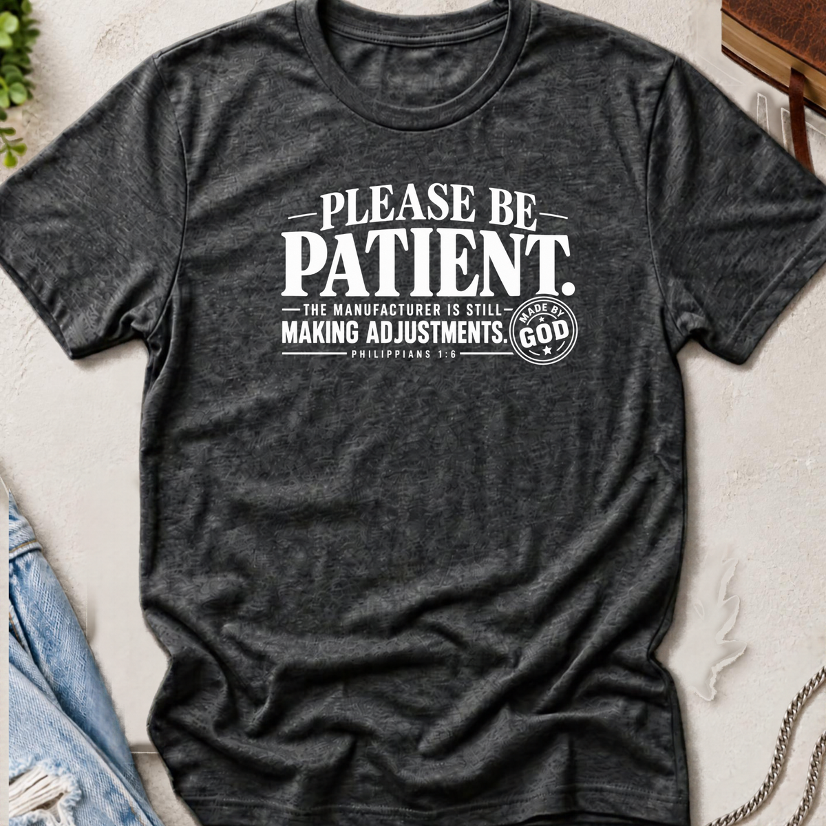 A dark heather gray graphic t-shirt laid flat on a textured light background, featuring the white printed text 'PLEASE BE PATIENT. THE MANUFACTURER IS STILL MAKING ADJUSTMENTS.