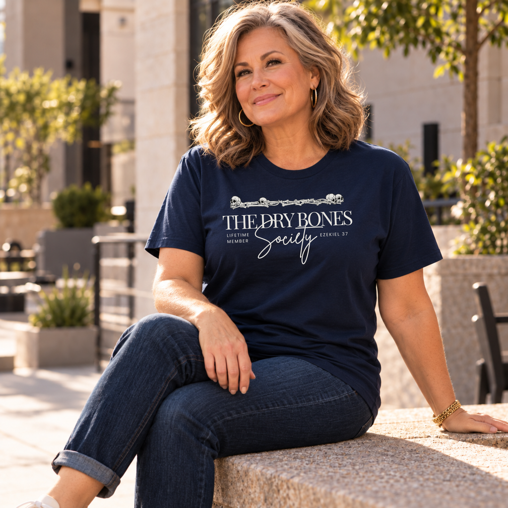 A woman sitting outdoors on a stone ledge wearing a navy blue graphic t-shirt with 'THE DRY BONES Society' printed in white, paired with dark blue jeans.