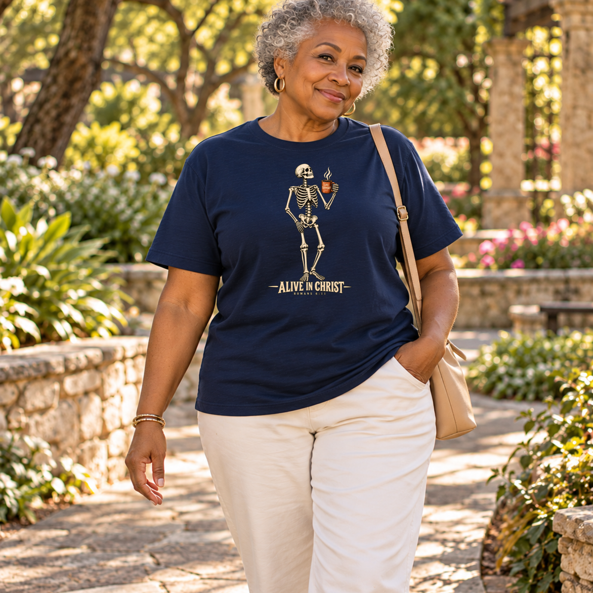 An older woman walking outdoors on a stone path wearing a navy blue graphic t-shirt with a skeleton holding a cup and the text 'ALIVE IN CHRIST', paired with off-white pants and a tan shoulder bag.