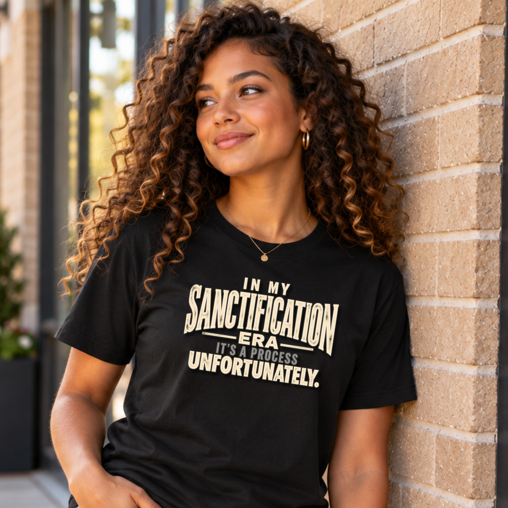 A woman with long, curly brown hair leans against a beige brick wall, wearing a black graphic t-shirt with the text 'IN MY SANCTIFICATION ERA IT'S A PROCESS UNFORTUNATELY.' printed in cream.