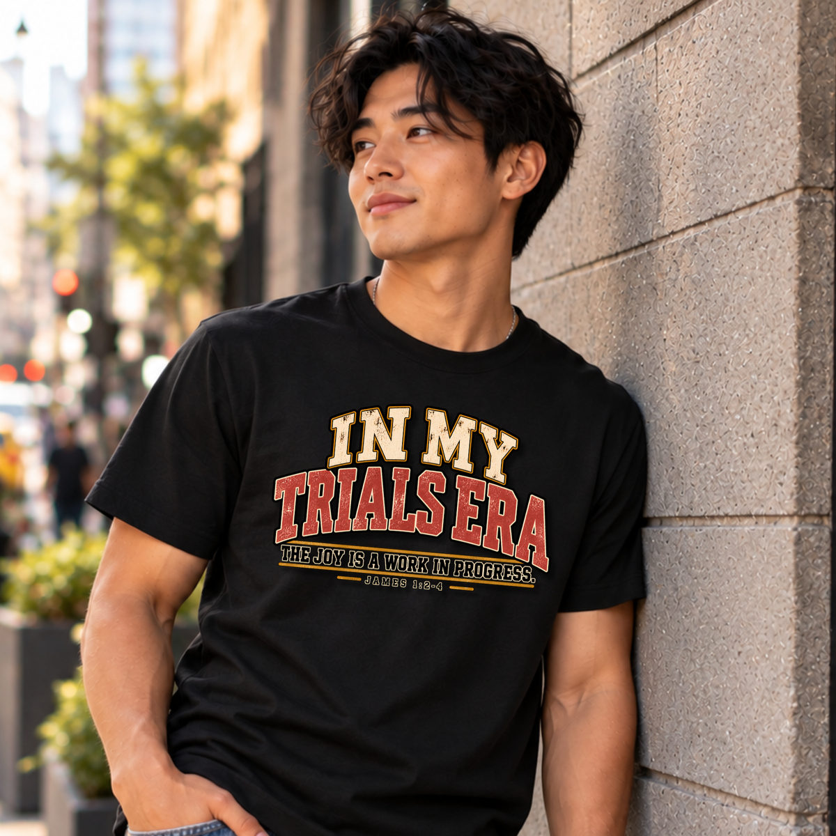 A young man leaning against a concrete wall looking out wearing a black christian t-shirt by SeedSowerGraphics  with typography that says "in my trials era' the joy is a work in progress. James 1:2-4 in cream, red, and black lettering