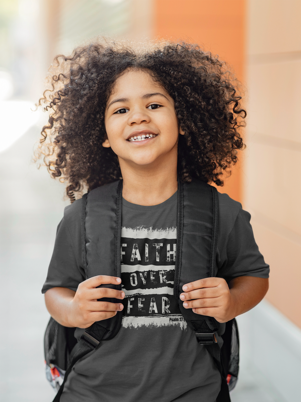A smiling young kid with voluminous curly hair stands outdoors wearing a black graphic T-shirt with the words 'FAITH OVER FEAR' and a black backpack.