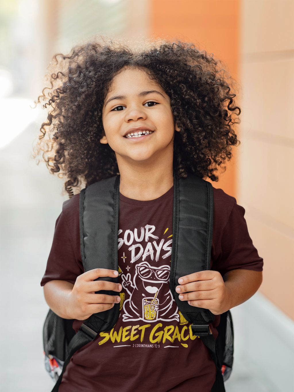 A smiling young child with curly hair wearing a maroon graphic t-shirt and a black backpack, standing outdoors against a blurred orange and beige background.