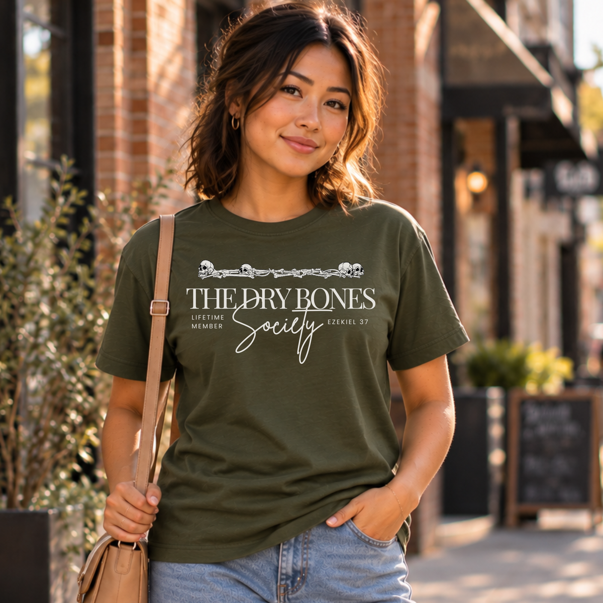 A woman wearing an olive green graphic t-shirt with 'THE DRY BONES Society' printed on the front, standing outdoors on a city sidewalk.