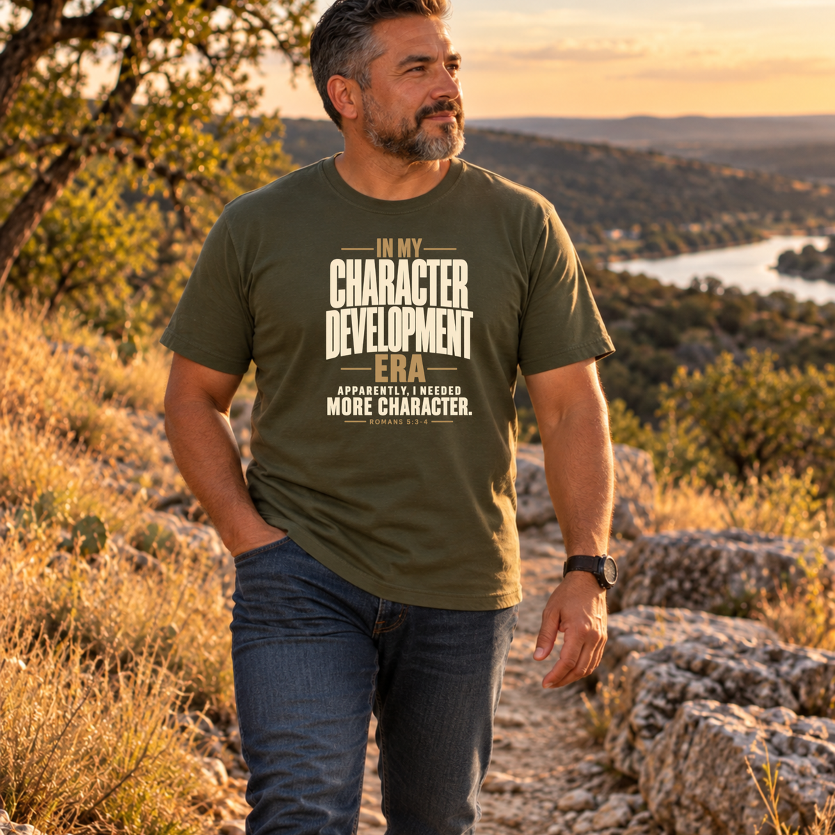 A man wearing a military green Christian graphic t-shirt by SeedSowerGraphics and blue jeans stands on a rocky outdoor trail at sunset, looking off-camera. The t-shirt features a distressed cream-colored text graphic that reads: 'IN MY CHARACTER DEVELOPMENT ERA Apparently, i needed MORE CHARACTER.