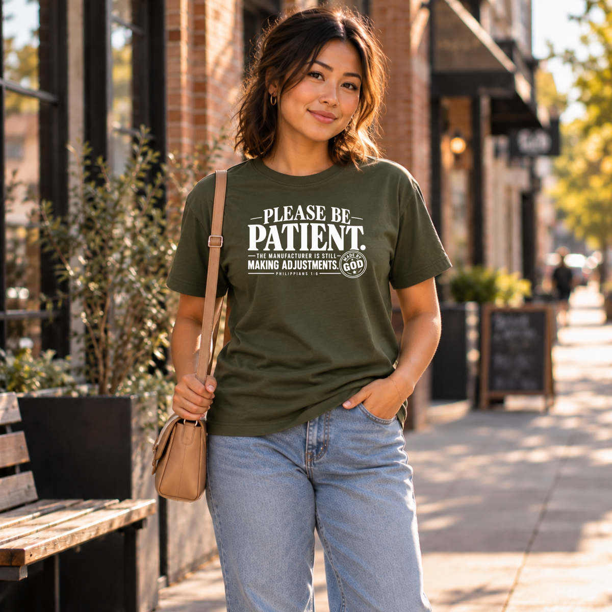 A woman stands on a city sidewalk wearing an olive green graphic T-shirt with white text that reads 'PLEASE BE PATIENT.