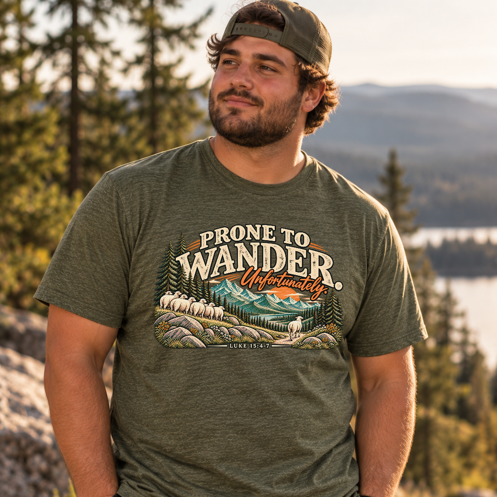A man standing outside wearing a heather military green Christian graphic t-shirt by SeedSowerGraphics. The t-shirt features a retro-style scenic graphic with sheep, mountains, and the text 'PRONE TO WANDER.Unfortunately. Luke 15:4-7