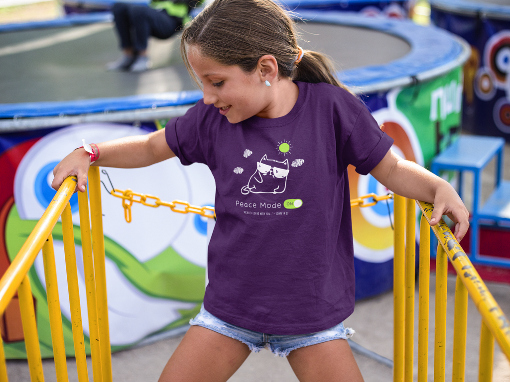 A young girl wearing a purple t-shirt with a 'Peace Mode' graphic stands at a yellow gate at an outdoor fair, holding the yellow metal bars.