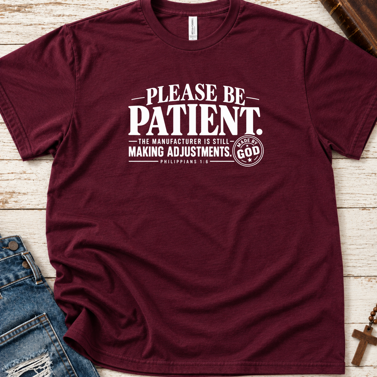 A maroon short-sleeve graphic T-shirt laid flat on a whitewashed wood surface, with blue jeans and a small cross visible as styling props. The shirt has large white text reading “PLEASE BE PATIENT.