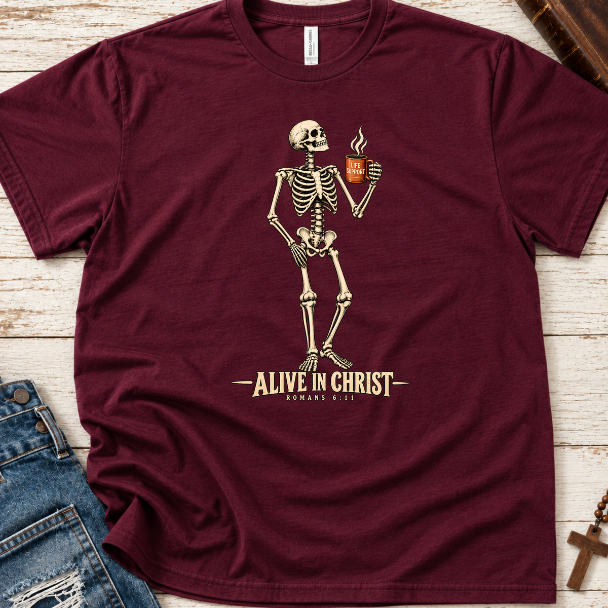 A maroon short-sleeve graphic T-shirt laid flat on a whitewashed wood surface, styled with blue jeans, a book, and a cross. The shirt features a cream skeleton holding a cup with steam, above the text 'ALIVE IN CHRIST' and 'ROMANS 8:11'.