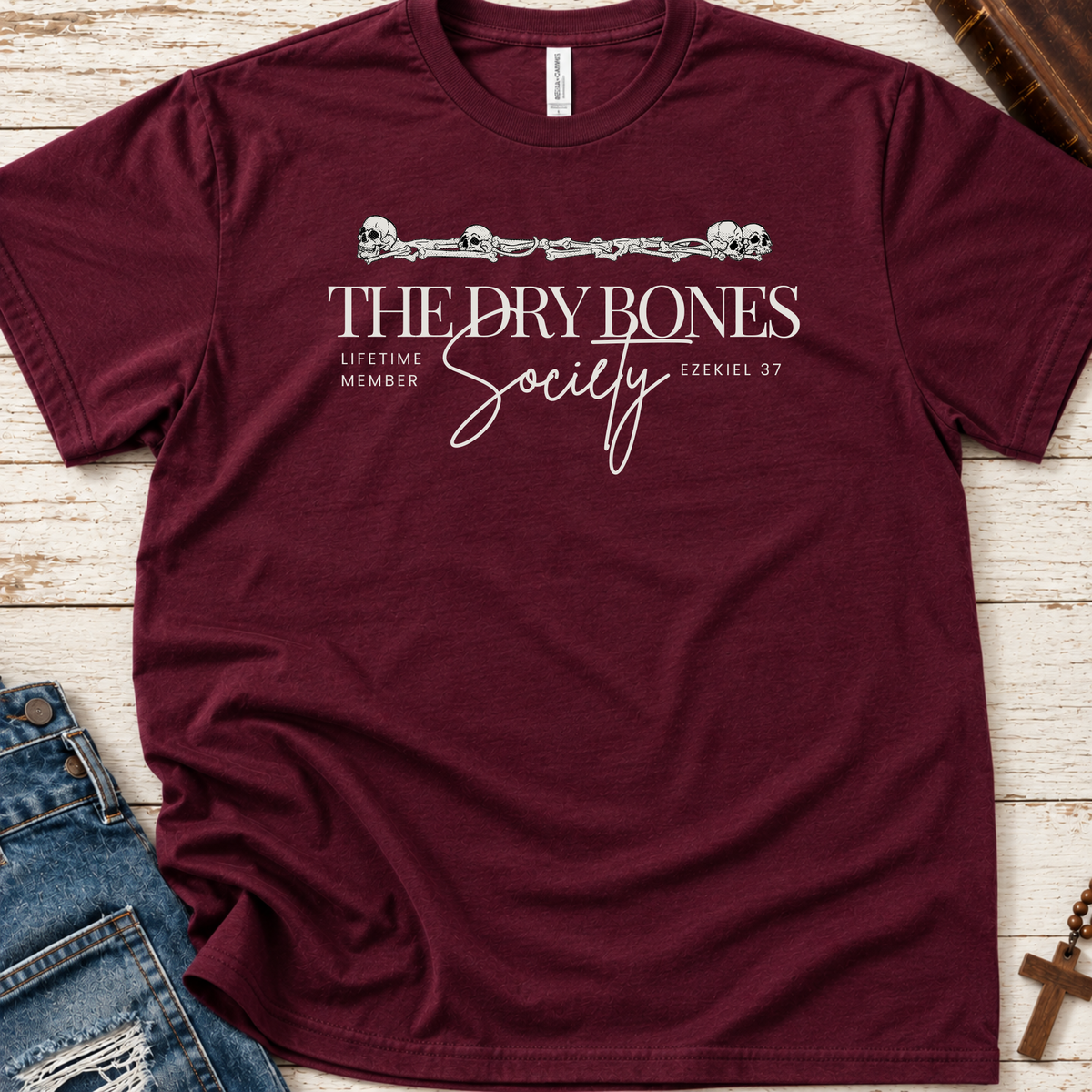 A maroon short-sleeve Christian graphic T-shirt by SeedSowerGraphics laid flat on a whitewashed wood surface, styled with blue jeans, a wooden cross, and a book corner. The shirt has a white printed design reading “THE DRY BONES Society” with smaller text “LIFETIME MEMBER” and “EZEKIEL 37,” plus a decorative barbed-wire and skull motif across the chest.